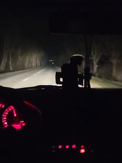 The image depicts a nighttime scene observed from the interior of a moving vehicle on a rural road near Koilovtsi, Bulgaria. The road, an asphalt surface with white dashed center markings, extends into the distance, illuminated by the vehicle's headlights. Dense, dark vegetation or trees line both sides of the road, partially lit by the vehicle's forward illumination. Several faint lights are visible in the far distance. Inside the vehicle, the lower-left section of the frame shows a portion of the dashboard, including an illuminated instrument cluster with a bright red glow. Below the center of the windshield, a dark, silhouetted object, possibly a dashboard accessory or phone holder, is visible. Further right and lower on the dashboard, a series of small, individually illuminated red buttons are present, one appearing to be a hazard warning light switch. The bottom edge of the vehicle's rearview mirror is visible at the top center. No occupants are directly visible.