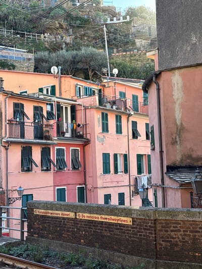 This image depicts a dense residential area in Vernazza, Italy, characterized by multi-story buildings constructed on a steep hillside. In the foreground, a dark brick wall with a weathered top surface spans horizontally across the lower frame. Affixed to the brick wall are two rectangular yellow signs with red borders and black text: "Vietato attraversare i binari" and "Do not cross the railway lines," indicating proximity to railway tracks, which are partially visible at the bottom left, showing two steel rails on ballast.
Immediately behind the brick wall and extending upwards are numerous tightly packed buildings, primarily painted in shades of peach, pink, and reddish-brown. These structures feature multiple windows fitted with dark green louvered shutters, many of which are open at various angles. Several balconies with black metal railings are visible on the facades, some displaying hanging laundry, including colorful garments on a line on a central balcony. Satellite dishes are mounted on the rooftops of some buildings. Signs of wear, such as faded paint and minor stucco damage, are present on building surfaces. A dark rain gutter system is visible on the rightmost building.
Above the buildings, a very steep hillside rises, densely covered with green vegetation, including small trees and shrubs. Terraced sections and retaining walls are carved into the hillside, with faint pathways and what appear to be distant structures, possibly smaller buildings or greenhouses, integrated into the landscape. Multiple overhead wires, likely power or utility lines, crisscross the upper portion of the frame, supported by utility poles. Two dark, ornate street lamps with glass enclosures are visible on a metal railing in the lower left and attached to a building facade on the right. No people are visible in the scene. The lighting suggests a clear day, with sunlight illuminating the buildings and hillside from the upper right.