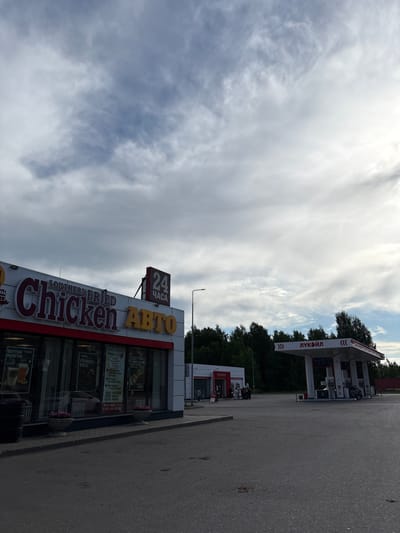 The image shows a Lukoil gas station and a Southern Fried Chicken restaurant in Krilovo, Russia. The restaurant has a sign indicating "24 hours" and "Auto" and advertisements for various menu items on its windows. The Lukoil gas station is visible in the background, beyond the trees behind the Southern Fried Chicken restaurant. The sky is cloudy. A paved road is visible in the foreground.