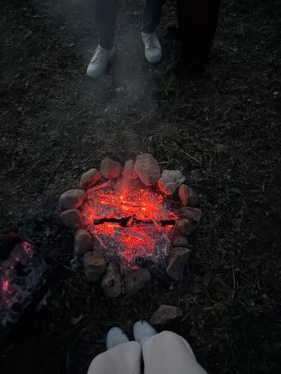 The image depicts a nighttime outdoor scene in Міхалішкаўскі сельскі Савет, Belarus, illuminated by two sources of fire. In the center, a circular fire pit is constructed from approximately 15-20 irregular, light-colored stones, containing a bed of intensely glowing red embers and partially charred wood. White ash is visible among the embers, and faint wisps of smoke rise from the fire pit.

In the lower-left corner, a smaller metal brazier or grill holds a separate collection of glowing red embers.

Three individuals are partially visible around the fires. At the top-left of the frame, a person wears dark trousers and light grey lace-up shoes. To the right of the frame, another individual wears dark clothing and dark, indistinct shoes. In the bottom-center foreground, a third person is positioned closest to the main fire, wearing light-colored (possibly white or light grey) casual trousers and matching slip-on sneakers, with their feet directed towards the embers. The ground consists of dark earth with sparse patches of grass and scattered leaf debris.