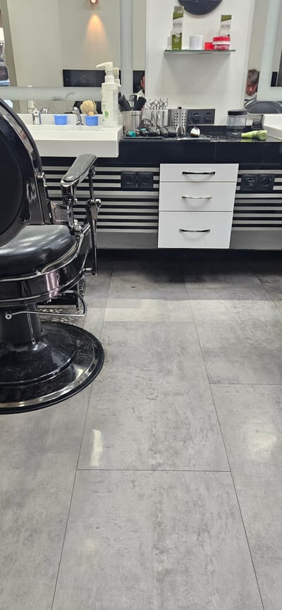 The image depicts a barber shop interior, likely in Gütersloh, Germany, given the location mentioned. The scene is captured from a high-angle perspective, showcasing a portion of the shop.
A black barber chair dominates the foreground, with its metallic components reflecting light. A barber station is positioned in the background. It features a white countertop, storage cabinets with silver handles, and various barber tools and products. A mirror reflects the barber, a man, and shop supplies. The background wall is adorned with black and white vertical stripes, incorporating electrical outlets.
The floor consists of large, light-gray stone tiles. Overall, the scene exudes a clean, modern aesthetic characteristic of a professional barber shop.