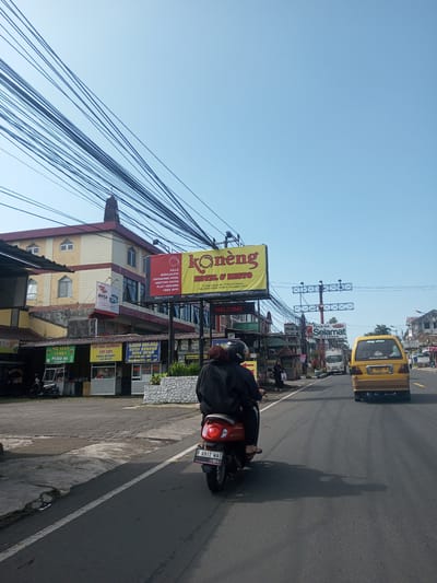 The image depicts a street scene, possibly in Puncak Cipanas, Indonesia, given the signage in the image and the OCR text. A person is riding a red scooter towards the camera. A yellow car is ahead in the same lane. On the left side of the street, there are food stalls with signs advertising "Mie Ayam Yamien Pedo 86" and "Sop Kaki Kambing Sop Sapi." A large billboard advertises "Koneng Hotel & Resto." A building is behind the billboard. Above the street, there are a mass of overhead power lines. The sky is clear.