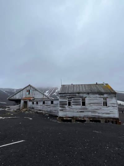 Two weathered, abandoned wooden buildings stand against a gray, overcast sky. The structures exhibit visible signs of decay with missing windows and deteriorated paint. They sit on a dark, grainy surface, possibly volcanic sand. In the background, mountains are partially obscured by fog or clouds, contributing to the overall desolate and remote atmosphere. The image is rotated ninety degrees clockwise.