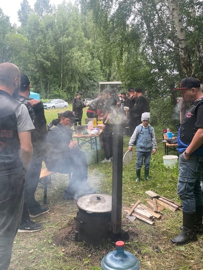A group of individuals, some wearing biker vests, are gathered outdoors in what appears to be a wooded area near Il'ichevskii, Russia. A large pot is being heated over an open fire, with smoke rising from it. A young child in a denim jacket and green boots stands nearby, holding a plate. A table with food and drinks is set up to the right, with several people interacting around it. The ground is covered in grass and fallen leaves, and trees form a dense background. Several cars are visible in the distance.