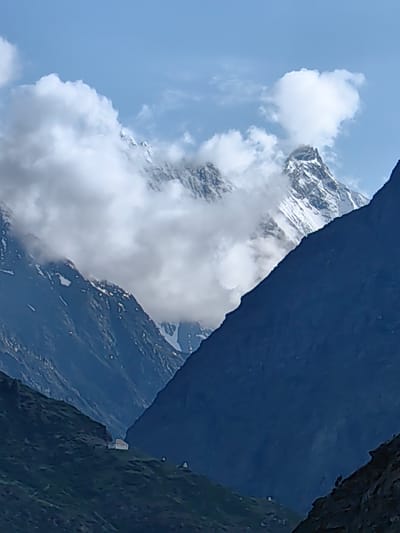 The image shows a dramatic mountain landscape with a clear blue sky and fluffy white clouds. In the foreground, dark, steep mountain slopes dominate the scene, with one slope on the right appearing almost entirely dark blue. To the left, a grassy, green slope is visible, dotted with a few small white structures, possibly buildings or shelters, on a ridge.

In the mid-ground, jagged, snow-capped mountain peaks pierce through a veil of white clouds. These peaks are stark and imposing, with visible rock faces and snow-covered ridges. The clouds partially obscure the mountains, creating a sense of depth and mystery.

The overall environment suggests a high-altitude mountainous region. While the description requests location in Kardang, India, there are no specific landmarks or textual information within the image to confirm this. The scene is devoid of any visible people or discernible actions. The image focuses solely on the natural grandeur of the mountain range.