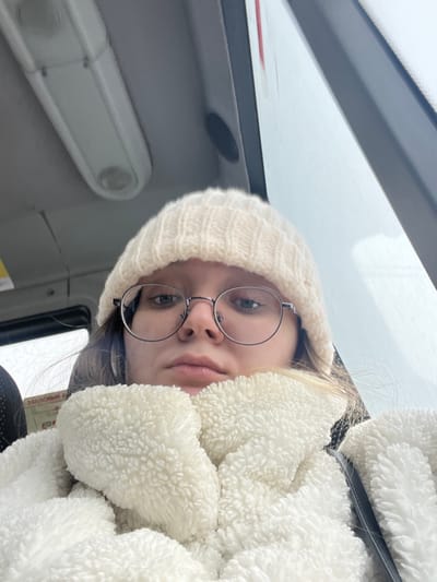 The scene is set inside a public transport vehicle, likely a bus or trolleybus, in the city of Борисово, Russia. A young individual, presenting as female, is captured in a close-up, front-facing perspective. She wears a cream-colored ribbed knit beanie and a cream-colored, textured fleece-like jacket that covers her neck and chest. Round, metal-framed glasses are worn over her eyes, and her hair is visible beneath the beanie. Her expression is neutral. The interior environment includes a light gray ceiling with a prominent white rectangular overhead light fixture. To the right of the subject, a window shows a bright, overcast exterior. To the left, a dark-colored seat back is partially visible, along with a white sign containing black Cyrillic text, partially legible as "ЗАПАСНЫЙ ВЫХ[ОД]" (Emergency Exit).