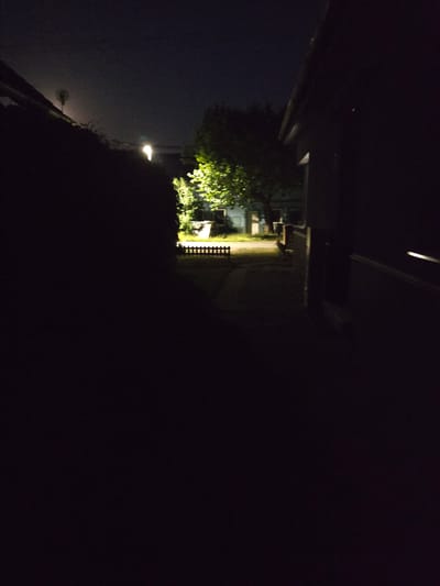 This image depicts a dimly lit outdoor scene at night, possibly an alleyway or backyard in Kostelec na Hané, Czechia. The left side of the frame is dominated by a dark, silhouetted hedge or shrubbery. To the right, a narrow pathway is visible, illuminated by a streetlamp that casts a bright pool of light in the distance. Further along the path, a cluster of trees is brightly lit, with some structures or buildings faintly visible behind them. A short, decorative wooden fence runs along the edge of the illuminated area. The right side of the image is largely obscured by the dark, shadowed side of a building. The sky above is dark, with a faint haze. There are no people or active events depicted in the scene.