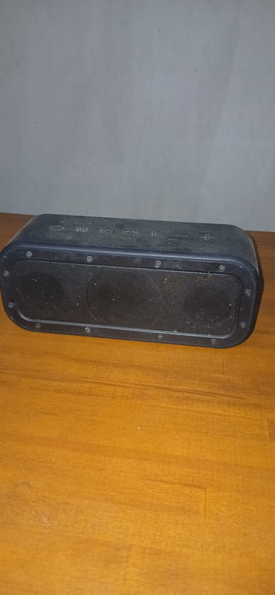 An indoor shot shows a black portable Bluetooth speaker resting on a light brown wooden table. The speaker is rectangular with a mesh grill on the front, covered by a black frame secured with visible screws. The top surface of the speaker has several buttons, including what appears to be a power button, an "M" button, an "EQ" button, and volume controls indicated by "+" and "-" symbols. The background is a plain, light gray wall. The image does not contain any elements that would indicate a specific location in Rokosovo, Ukraine, or any city.