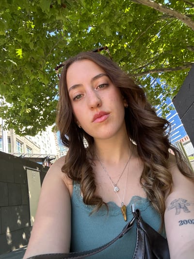 A young woman poses outdoors with green leaves and city buildings in the background. She is wearing a green top, two necklaces, and has a tattoo on her upper arm. Her brown hair is styled in loose waves. The photo is taken from a low angle, capturing her face with a slight tilt. Sunglasses are perched on her head. A black bag strap is visible on her shoulder. The lighting suggests it is daytime.