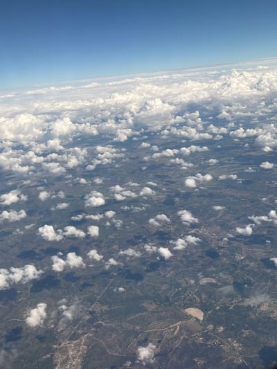 The image shows an aerial view of a landscape, with a focus on the transition between the sky/atmosphere and the terrain below. The sky is visible in the top left of the frame, showcasing a clear gradient from deep blue to a lighter, hazier blue near the horizon. Scattered, puffy white clouds partially obscure the view of the ground below, creating a dappled pattern of light and shadow.
The terrain appears to be rural, with a mixture of green vegetation, possibly forests or fields, interspersed with what seem to be bare patches or cultivated areas. The patterns suggest varying land use and natural features, potentially including small water bodies or drainage systems.
While there is no specific reference to the city of Coruche, Portugal within the image, the landscape type is representative of the general area. The image does not have any people, so there are no interactions or actions. It also does not show any objects or subjects except for the features listed.