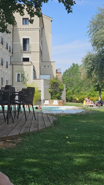 This media file captures a sunny day scene at what appears to be a resort or hotel in Alhama De Aragón, Spain. The primary focus is on an outdoor swimming pool area.
In the foreground, a wooden deck leads to the pool. Several black plastic chairs are arranged on the deck, some facing the pool and others angled away. The pool itself is visible, with tiled edges and clear blue water. Two people are partially submerged in the pool, with their heads and shoulders visible. One person, lying on their back, appears relaxed.
Beyond the pool, a well-maintained lawn stretches out, bordered by lush green trees. In the middle ground, several people are relaxing on lounge chairs on the grass. Some are wearing swimwear and appear to be sunbathing, indicating it's a warm day.
A large, multi-story building with a light-colored facade is situated to the left of the scene. The building has multiple windows, some with dark panes and others with metal gratings. Architectural details, such as decorative elements along the facade and balconies, are visible. Trees with dense foliage frame the building and extend towards the top of the frame.
The sky is a clear, bright blue, with a few wispy clouds. The sunlight suggests it is daytime, likely late morning or afternoon, given the shadows cast by the trees and the intensity of the light. The overall atmosphere is one of leisure and relaxation in a pleasant outdoor environment. No text is visible in the image.