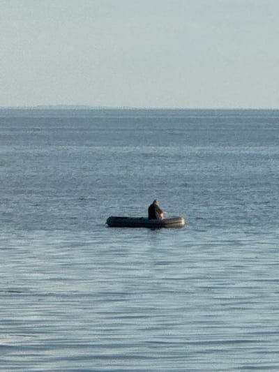 A person is sitting in an inflatable raft on a body of water. The raft is dark in color and appears to be a dinghy. The person is wearing dark clothing and is facing away from the viewer. The water is calm with visible ripples. The sky is a pale blue and appears to be overcast. The scene is likely a coastal area, given the calm water and the vastness of the horizon. The location is Shepelevo, Russia.