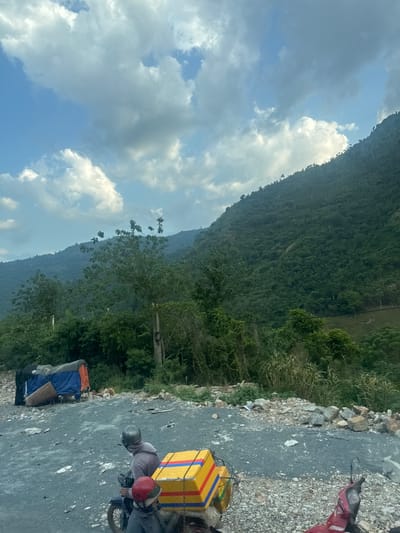 The image captures a scene on a road near Aê Thi, Vietnam, characterized by a mountainous backdrop and a partially paved surface. In the foreground, two individuals are seated on a motorcycle, both wearing helmets. The rear passenger is positioned behind the driver and carries a large, brightly colored yellow box with red and blue stripes secured to the back of the motorcycle. The motorcycle itself is dark in color. To the left, a makeshift shelter is visible, constructed with a blue tarp and an orange tarp, with corrugated metal sheets leaning against it. The ground is a mix of gravel and dark asphalt. The background features a dense, green mountainside covered in trees, with a cloudy sky above. The overall impression is one of a rural, potentially remote location.