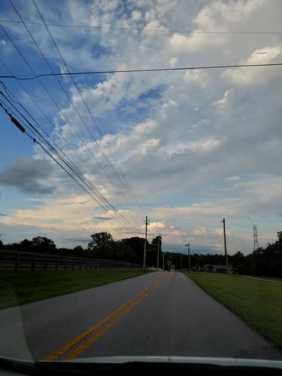 The image shows a paved road with double yellow lines, indicating a two-way street. The road is lined with grass on both sides. On the left side of the road, there is a wooden fence and a line of trees. On the right side, there are also trees, grass, and utility poles with power lines. In the distance, there are houses and a tall transmission tower. The sky is partly cloudy with a mix of blue sky and white and gray clouds. The scene appears to be a rural or semi-rural area in Wilmore, United States. There are no people or vehicles visible on the road, and no apparent action is taking place. The lighting suggests it is daytime, possibly late afternoon or early evening due to the diffused light and cloud cover.