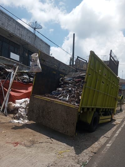 The image depicts a green dump truck with its bed raised, overflowing with scrap metal and debris. The truck is parked on the side of a street, with some of the scrap metal spilling onto the pavement. A bag hangs near the front of the truck bed. Behind the truck, there is a building with a satellite dish on its roof. The sky is partly cloudy. A person is standing on the sidewalk near the back of the truck. Based on image similarity, the location may be Kioumaknao 2, Laos.