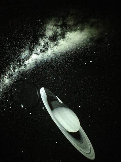 This is a close-up, angled shot of a model or projection of Saturn and a segment of the Milky Way, set against a black background that simulates space. The planet Saturn is prominently featured in the lower right quadrant of the image, depicted with its characteristic rings. The rings are layered and appear to be made of a lighter material than the planet itself, with visible segmentation and subtle variations in tone. The planet's surface is also visible, with a darker, textured upper portion and a lighter, smoother spherical body.
To the upper left of Saturn, a nebulous band of light, representing the Milky Way, stretches diagonally across the image. This band is comprised of a multitude of tiny white dots, representing stars, and a more diffuse, luminous cloud-like area. The black background is sparsely populated with additional white dots of varying sizes, further enhancing the illusion of a starry expanse. A dark, circular shadow or object is visible to the left of Saturn, possibly representing another celestial body or a lens artifact. The lighting appears to be coming from the upper left, illuminating the Milky Way and casting subtle shadows on Saturn.
The image does not contain any people, actions, or interactions. It is a still depiction of astronomical objects. There is no indication of a specific location in Jiguan District, China, as the scene is entirely artificial and represents outer space.