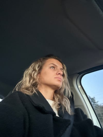 A young woman with wavy blonde hair and fair skin is looking upward, presumably out of the sunroof of a car. She is wearing a black coat over a white top. The view outside the sunroof shows a blue sky and a glimpse of trees. The interior of the car is mostly dark, with the roof dominating the left side of the frame.
