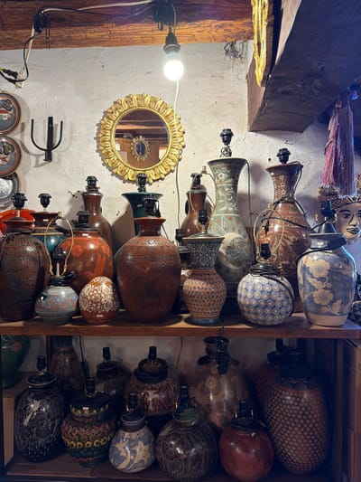 This image captures an indoor scene, likely a workshop, retail space, or storage area for decorative items, particularly lamp bases. The photo is oriented vertically, with what would normally be the left side of a room becoming the top, and the floor/shelving area becoming the right side.
The primary focus of the image is a dense collection of ceramic or pottery lamp bases, displaying a remarkable variety of shapes, sizes, colors, and intricate patterns. These lamp bases are arranged in several rows along what appears to be a wall display on the left side of the frame (when viewed in correct orientation) and packed into a wooden shelving unit on the right side.
On the left side of the image (which would be the upper wall and ceiling in a correctly oriented view), a bright, exposed incandescent light bulb hangs from a wire, providing direct illumination to the scene. Below it, a circular mirror with a ornate, golden, sunburst-like frame is mounted on the textured white wall. A reflection of another smaller mirror is visible within it. Further down this wall, a black metal decorative hook or sconce is attached. In the bottom left corner, several round pottery plates with painted designs are partially visible.
The lamp bases themselves are diverse:
* Some are tall and slender, reminiscent of traditional vases.
* Others are more squat, globular, or cylindrical.
* Colors range from blues, greens, browns, and reddish-orange hues to lighter cream and off-white tones.
* The patterns include elaborate floral motifs, geometric designs (dots, concentric circles, diamond patterns, zigzags), textured surfaces, and sometimes abstract or organic shapes.
* Each lamp base is fitted with a black electrical socket at its top, often with black wires trailing, indicating their function as lamp components.
To the right side of the image (which would be lower or floor-level shelving), a wooden shelving unit is filled with more of these decorative lamp bases. They are closely packed, some standing upright, many lying on their sides, showcasing an even greater abundance of designs and textures.
In the top left corner of the image (which would be the upper left wall/ceiling corner), several decorative items are visible:
* A cluster of colorful fabric tassels or fringes in shades of red, pink, and purple hangs from a wooden beam.
* Next to them, a stylized decorative mask or head, possibly made of wood or papier-mâché, with painted features, is also present.
Exposed rough-hewn wooden beams traverse parts of the background, suggesting structural elements of the building. The walls are an off-white or light gray color with a visibly rough, textured finish, possibly stucco or plaster. The overall lighting is bright and direct from the visible bulb, creating some shadows and highlights on the numerous objects. No people or visible text are present in the image. The scene offers a rich visual catalog of artistic pottery and lamp designs, hinting at a place of craftsmanship or retail for unique home decor.
