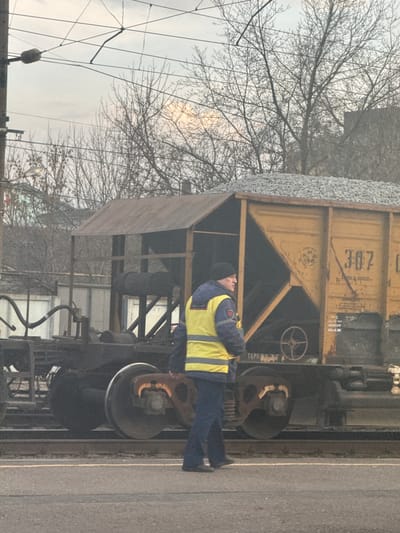 An outdoor scene depicts a rail worker inspecting a railcar, likely in or near Gryazi, Russia. The railcar is a yellow hopper car loaded with what appears to be gravel or ballast. The worker is wearing a dark jacket under a yellow safety vest and dark pants. He is standing next to the railcar's wheels, looking upwards towards the car's body. The environment includes overhead power lines, bare trees indicating winter or early spring, and what may be a building in the background. The lighting suggests an overcast day.
