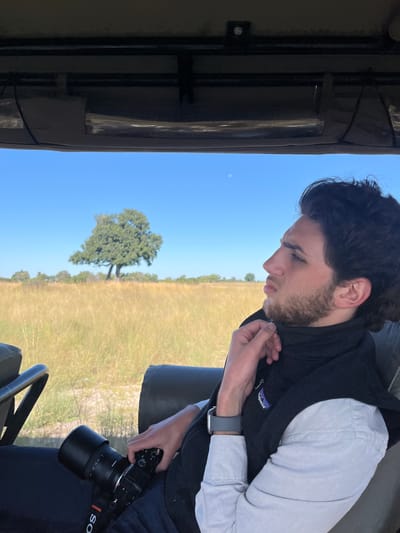 A man is seated in an open-top vehicle, possibly a safari vehicle, in Kakoro, Botswana. He is wearing a light-colored shirt, a dark-colored vest with a visible Patagonia logo, and an Apple Watch. He is holding a black Sony camera with a large lens. Outside the vehicle, the landscape is a grassy field with a single tree in the distance under a clear blue sky. Part of the vehicle's frame and roof is visible at the left edge of the image. The man appears to be adjusting the collar of his shirt or vest.