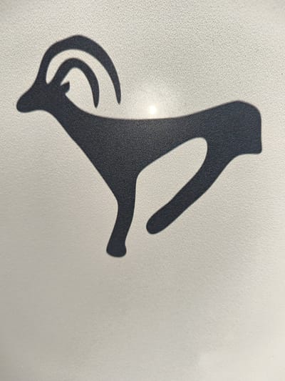 The image shows a dark silhouette of a stylized ibex or mountain goat with large curved horns against a light-colored, textured background. The silhouette is depicted in profile, facing left, with its body elongated and its legs bent as if in motion. A bright light source is visible behind the head and horns of the animal, creating a subtle halo effect. The background appears to be a textured wall or surface. There are no people or specific landmarks visible in the image. The location in Mayma, Russia is not identifiable from the image content.