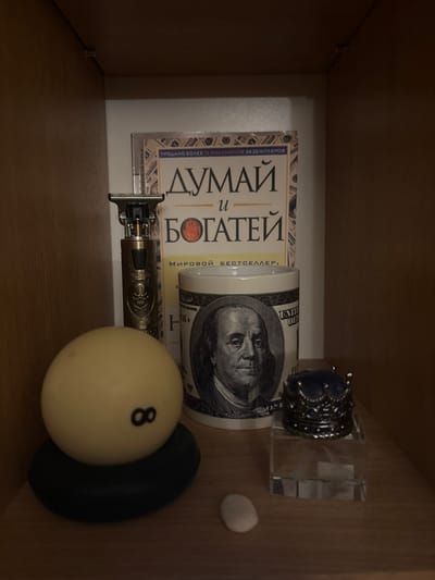 A collection of items is displayed on a wooden shelf. From left to right, there is a pool ball with the number "8" on it, resting on a dark, circular base. Next to it is a metallic electric trimmer with a skull design. Behind the trimmer and pool ball, a book with a golden cover and Cyrillic text is partially visible. To the right of the book is a white mug featuring the portrait of Benjamin Franklin and dollar bill designs. In front of the mug, a small, transparent perfume bottle with a decorative crown-shaped cap is placed. To the right of the pool ball and slightly in front of the mug, a small, light-colored stone is visible on the shelf. The background appears to be the interior of a wooden cabinet or shelf. The lighting is dim, casting shadows on the objects.