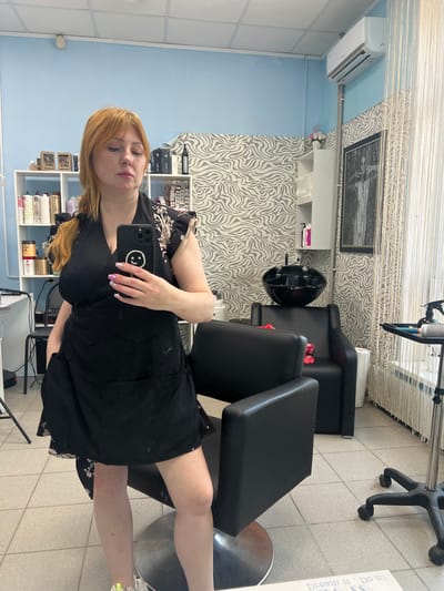 A selfie photograph shows a woman seated in what appears to be a hair salon in Shuya, Russia. The woman has red hair and is wearing a black garment, holding a smartphone to take the picture. Her manicure is visible. The background includes salon equipment: a shampoo bowl, black chairs, a rolling chair, and shelves stocked with products. The walls are painted in blue and have a zebra-striped wallpaper. Blinds are hanging from the ceiling. An air conditioner is mounted on the wall. A framed picture is present.