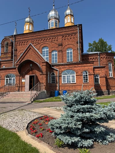 The image depicts the exterior of an orthodox church constructed from red brick in the city of Chudovo, Russia. The church features multiple domes topped with crosses, arched windows, and an entrance with a covered portico. In the foreground, a landscaped area with a small blue spruce tree and red flowers is visible. The sky is clear and blue. There are no people or animals present in the image.