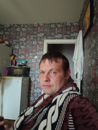 The image is a selfie taken indoors, likely in a residential setting. A man with short brown hair and blue eyes is the central subject, wearing a patterned robe. He is positioned in the foreground, with a portion of his face and upper body visible. The background features wallpaper with a repeating pattern of coffee-themed text and imagery. To the left of the man, a refrigerator and a microwave are visible, suggesting a kitchen area. A door is present in the background, and a white towel or cloth is hanging on it. The lighting appears to be artificial, casting a slight shadow on the man's face. The overall impression is that of a casual, personal moment captured in a domestic environment. No specific landmarks or indicators place this scene in Андрэеўшчынскі сельскі Савет, Belarus.