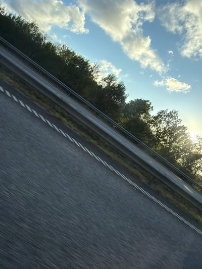 A tilted, close-up shot captures a highway's asphalt lane in Ödsmål, Sweden. The lane is marked with white dashed lines and appears blurred due to motion. A silver guardrail runs parallel to the lane, separating it from a grassy verge. Beyond the verge, a dense line of green trees flanks the road. The sky above is a bright blue, dotted with fluffy white clouds illuminated by the sun, which is peeking through the trees on the right side of the frame, casting a warm glow. The perspective suggests the photo was taken from a moving vehicle.
