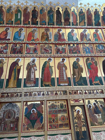 The image displays a section of an iconostasis, a wall of icons and religious imagery found in Orthodox churches. This particular iconostasis is elaborate, featuring multiple rows of painted icons. The icons are depicted on gilded backgrounds and portray religious figures, saints, and biblical scenes in a traditional Byzantine style. The figures are depicted in stylized robes with halos, and many hold scrolls or religious artifacts. The colors are rich and vibrant, with prominent use of gold, red, blue, and green. The overall scene is one of religious devotion and artistic tradition. This iconostasis is located in the Solovetskiy Monastery, on Solovetsky Island in the Arkhangelsk Oblast of Russia.