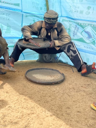 An individual wearing a checkered head covering and jacket, dark pants with a skull design, and sandals is squatting on a surface covered with what appears to be light blue bags with agricultural logos. They are holding a flat, round object with both hands. Next to them on the sandy ground is another similar round object. The setting is outdoors, with a sandy ground surface. The location is Maiyanga, Nigeria.
