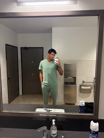 The image shows a reflection in a mirror in what appears to be a public restroom. A man in light green scrubs is taking a selfie. Behind him are gray restroom stalls and a paper towel dispenser. On the countertop next to the mirror are a faucet, soap dispenser, and cleaning products. Based on contextual information and the cleaning product visible, the restroom is located in El Segundo, California, USA.