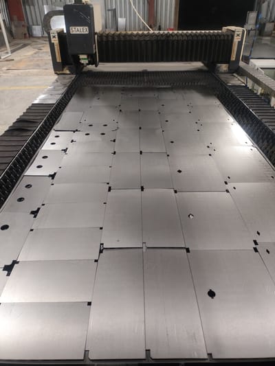 An overhead, wide-angle shot captures a large, industrial laser cutting machine with a metal bed covered in sheets of metal. The machine's cutting head, labeled "STALEX", is visible at the top left of the frame, positioned above the metal sheets. The metal bed is a grid of interlocking pieces, with some sheets showing pre-cut shapes and circular holes. The environment appears to be an indoor workshop or factory, with concrete flooring and industrial structures in the background. The lighting suggests an indoor, well-lit space. This scene is located in the city of Dangir, Uzbekistan, within a manufacturing facility.