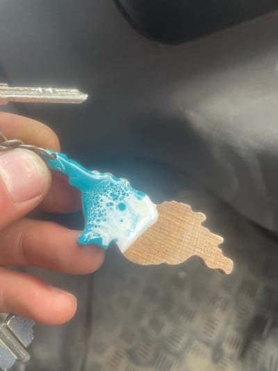 The image does not depict a location in the city of Juta, Georgia. The scene is a close-up, depicting a human hand holding a keyring assembly. The hand's skin appears light-toned, with visible fingerprints and clean fingernails on the thumb, index, and middle fingers. The keyring includes at least two metallic, silver-colored keys, with one partially visible at the upper left and another at the lower left. The primary object is a decorative keychain attached to the keys via a metal split ring. This keychain is shaped like a stylized landmass or island, approximately two-thirds of which is covered in a turquoise-blue and white epoxy resin, exhibiting a detailed cellular or "sea foam" pattern. The remaining one-third of the keychain is made of light-brown wood, displaying distinct vertical grain lines. The background is dark and out of focus, indicating an interior setting, likely a vehicle. Visible elements of the background include a dark gray, smooth plastic surface in the upper portion and a dark, textured surface resembling a rubber or metallic diamond plate floor mat in the lower portion.