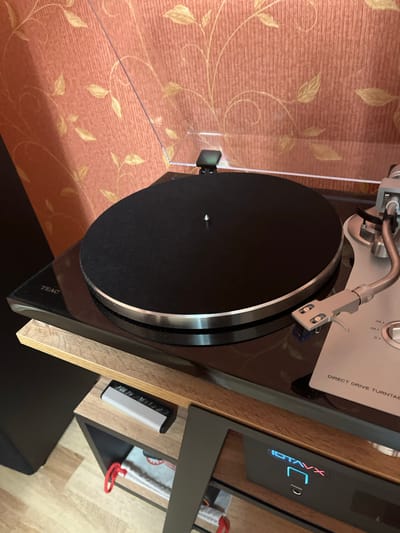The image shows a close-up of a turntable with a black platter and silver accents. The turntable is placed on a glossy black surface, possibly the top of a component rack. To the left of the turntable, the "TEAC" logo is visible. A tonearm with a silver cartridge is positioned over the platter. The background features a textured wallpaper with a repeating pattern of light-colored leaves on a reddish-orange surface. Below the turntable, a wooden shelf is visible, on which a remote control and a piece of fabric with a red loop are placed. Further down, a black electronic device with illuminated "IDTAVX" and "PHONES" branding is visible. The flooring is a light-colored wood laminate. The scene appears to be an indoor setting, likely a living room or entertainment area. No people are present in the image. The location is identified as Ivanovo-Grushevsky, Russia.