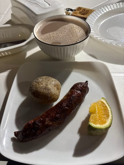The image captures an overhead, close-up view of a meal set on a light-colored surface, stated to be located in Quimbaya, Colombia. Dominating the foreground is a rectangular white ceramic plate presenting three distinct food items. On the left, a single, whole, unpeeled potato, light brown with visible textured skin, rests. To its right, an elongated, dark brown sausage, appearing cooked and slightly charred, is positioned horizontally across the plate. On the far right of the plate, a wedge of a citrus fruit displays vibrant orange pulp and a section of green rind, with a small portion seemingly bitten or cut from its top edge.

Behind the main plate, a white bowl with a ridged exterior holds a light brown, frothy beverage, consistent with hot chocolate or a similar warm drink. To the left of the bowl, a white, possibly plastic or ceramic, caddy or dispenser is partially visible, containing what appears to be utensils or napkins. To the right of the bowl, a stack of white, fluted-edge disposable plates is present. Further in the background, a small, light-colored object, possibly a wooden keychain or charm, lies on the surface. The scene is illuminated by artificial light, indicated by pronounced shadows cast by the objects onto the table, suggesting an indoor setting or an outdoor area during low light conditions. No individuals are visible within the frame.