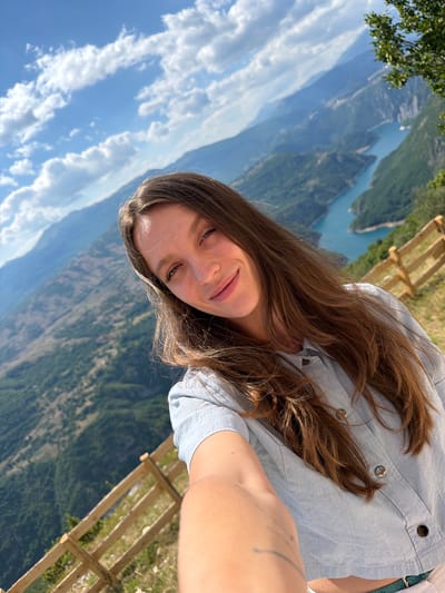 A young woman with long brown hair takes a selfie from a scenic overlook. She is wearing a light blue collared shirt. Her left arm is extended, holding the camera. Behind her, a winding turquoise river flows through a lush green mountainous valley under a blue sky with white clouds. A wooden fence runs along the edge of the overlook. The location is the Bukovac viewpoint in Montenegro, which offers panoramic views of the Piva River Canyon.