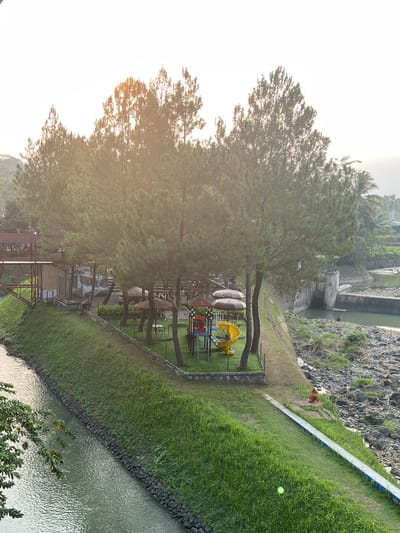 The image captures a waterside recreational area in Krasak, Indonesia, bathed in the soft, warm light of what appears to be late afternoon or early morning. The scene is dominated by tall, slender pine trees that cast long shadows across the meticulously manicured green lawn.
In the foreground, a canal or river flows gently, its surface reflecting the overcast sky. The bank of the canal is reinforced with dark, rough-hewn stones and covered in lush green grass. A narrow, paved pathway, bordered by a blue stripe, runs alongside the canal, leading towards the right of the frame.
Further back, on a slightly elevated grassy terrace, a small children's playground is set up. It features a yellow slide, a red climbing structure, and a blue platform. Scattered around the playground are a few tables and chairs, some partially covered by thatched umbrellas, suggesting an outdoor seating area for a cafe or restaurant.
To the left of the playground, a rustic wooden bridge or walkway extends over the canal, connecting to a structure with a red roof and bamboo walls, possibly a building or an elevated dining area.
In the background, a concrete dam or retaining wall rises from the rocky bank of a wider body of water. The surrounding landscape is dotted with more trees, including palm trees, and hints of distant buildings can be seen. A solitary figure is visible near the dam, seemingly fishing in the water. The overall impression is one of a tranquil, well-maintained park designed for leisure and family enjoyment.