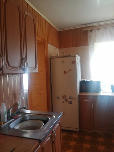 The image depicts a domestic kitchen scene, likely in Dubrovka, Russia. The setting is characterized by wooden paneling on the walls and a tiled backsplash above the countertops. In the foreground, a stainless steel sink is visible, with a partially filled dish inside and a faucet above. To the left of the sink, a bottle of liquid soap is present.
Dominating the mid-ground is a tall, white refrigerator adorned with several colorful stickers. The brand name "Indesit" is clearly visible on the upper part of the refrigerator door, along with a circular logo. To the right of the refrigerator, a dark-colored microwave oven sits atop a wooden cabinet. This cabinet, along with another to its right, features wooden doors with simple handles. Above the refrigerator and cabinets, a sheer white curtain covers a window, allowing a bright, possibly daylight, illumination into the room.
In the background, to the left of the refrigerator, a doorway is visible, leading to another area that appears to be lit. Overhead, the ceiling is a light color, possibly white or a pale shade. The flooring is a patterned linoleum or laminate in shades of brown. The overall impression is of a functional, lived-in kitchen. There is no discernible activity taking place, and the image captures a static moment. The lighting suggests it could be daytime, with sunlight streaming through the window.