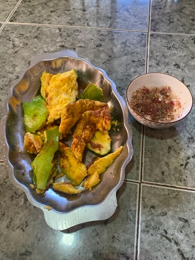 The image captures an indoor scene in Đak Đoa, Vietnam, displaying food items arranged on a tiled floor. A metallic tray with a scalloped edge, possibly stainless steel, occupies the left side of the frame. Inside the tray are various pieces of what appears to be ripe avocado, some retaining sections of green skin, and other yellowish pieces, possibly cooked egg or very ripe avocado flesh. A reddish-brown spice blend is sprinkled over many of these items. To the right of the tray, a small, white ceramic bowl with a subtle decorative pattern and a brown rim contains a granular condiment composed of white, dark, and reddish-pink particles. The surface supporting these items is a floor covered with dark grey or green-grey square tiles, arranged in a grid pattern with visible grout lines. No individuals are present.