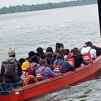 A red motorized boat transports approximately 18 to 20 individuals across a body of water in Koko, Nigeria. The vessel is an open-top boat, generating a visible wake, indicating movement. Most passengers are adults, and a minimum of 15 are wearing orange life jackets, several of which display "DEAN MARINE" text on the back. One person at the far left (stern) wears a light-colored beanie and a dark backpack over a grey hooded garment. Another individual is partially visible in a yellow hooded garment. An orange cylindrical object, likely a fender or lifebuoy, is secured inside the boat. The water is greyish-green and rippled. The distant background features a dense, dark green treeline, indicative of land, beneath an overcast, light grey sky.