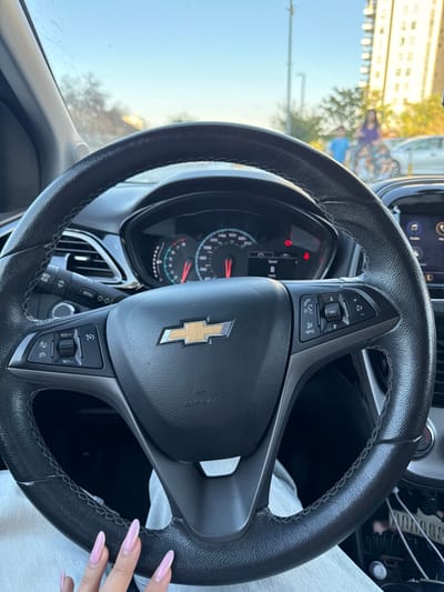 The image depicts the interior view from a stationary Chevrolet vehicle, looking forward through the windshield into a daytime urban or suburban environment within Ganei Tikva, Israel.
In the foreground, a black leather-wrapped steering wheel with a gold Chevrolet emblem is prominent. The steering wheel features multi-function controls on both sides: cruise control functions ("RES+", "SET-", "CANCEL") on the left, and audio/call controls (phone, source/seek, volume, voice command) on the right. A hand with long, light pink acrylic nails is visible resting on the lower left portion of the steering wheel, wearing light-colored trousers.
Behind the steering wheel, the instrument cluster is visible, indicating a speed of 0 km/h. Several warning lights are illuminated, including a parking brake indicator and what appears to be an open door or seatbelt warning. A partial view of an infotainment screen, displaying icons such as "Audio," is visible on the right side of the dashboard.
Outside the vehicle, the sky is clear and light blue. In the mid-ground, slightly blurred, two individuals are visible: an adult figure in a purple top near a bicycle equipped with a child seat, which appears to be occupied by a small child. Another child, wearing a blue top, stands nearby. Further in the background, a multi-story light-colored apartment building stands on the right. A light-colored sedan is partially visible behind the figures. Vegetation and utility poles are also present in the background.