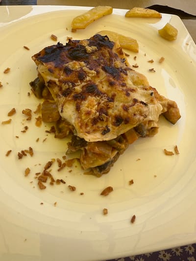 This image is a top-down, close-up shot of a meal on a light yellow, round plate. The primary subject is a portion of baked food, possibly a casserole or gratin, dominating the center of the plate. The food has a golden-brown, baked crust, with some areas appearing slightly overcooked or charred, particularly along the left edge. Visible beneath the crust are layers of vegetables and possibly cheese.

Scattered around the main dish are several thick-cut French fries, with a few positioned on the left side of the plate. Small, crispy fried onion bits are also sprinkled across the plate, adding texture and visual interest. The plate itself has a subtle, textured surface.

The lighting suggests it might be daytime, with natural light illuminating the food. The overall impression is that of a casual meal, perhaps taken at a restaurant or during a home-cooked meal. There are no people visible in the image. The setting appears to be a table, with a hint of a patterned tablecloth visible at the far right edge of the frame. The image does not contain any visible text.