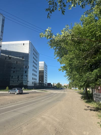 The image depicts a street scene in Kalinin, Russia, under a clear, bright blue sky, suggesting a sunny day. Lush green trees with dense foliage frame the top left of the image, casting dappled shadows. To the right, a paved road runs diagonally, with a gravel shoulder. Along the left side of the road, modern buildings with reflective glass facades dominate the scene. These multi-story structures have a contemporary architectural style, with numerous rectangular windows. A silver car is parked on the gravel shoulder near one of the buildings. Further down the road, more buildings are visible, some appearing residential. A large sign with red and white lettering is affixed to a tree on the right side of the road, though the text is partially obscured and difficult to read clearly. The overall impression is one of an urban or semi-urban environment on a pleasant day, with a contrast between the natural elements of the trees and the modern architecture.