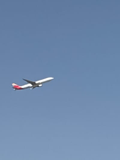 The image shows a commercial airplane with red and white markings on its tail, seen from below against a clear blue sky. The airplane appears to be in a steep descent, with its nose angled downwards. The sun is high in the sky, casting a bright light on the aircraft. The scene is set in an open area, likely above an urban or semi-urban environment, given the implied location of Coslada, Spain, which is near Madrid. There is no visible text in the image. The overall impression is one of an aircraft in distress, but without further context, the specific event or activity taking place remains uncertain.