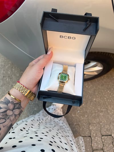 A person's hand, with manicured nails and gold bracelets, holds open a black gift box containing a gold-banded watch with a green face. The watch rests on a white cushion inside the box. The box has the "BCBG" logo printed in white. In the background, the edge of a silver car and a paved surface are visible. The scene is outdoors.