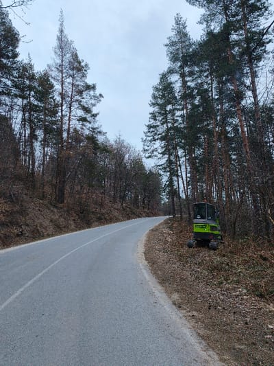 The scene depicts an unpopulated, forested roadway located in Sveti Konstantin, Bulgaria, under a uniformly overcast sky. A grey asphalt road, marked with a single solid white center line, curves gently to the right and extends into the distance. The road surface appears dry.
Dense forest flanks both sides of the roadway. On the left, the terrain slopes upward, covered primarily by bare deciduous trees and some conifers, with dry leaves and sparse undergrowth on the forest floor. On the right, an unpaved, earthen shoulder separates the road from a dense stand of tall pine trees and some bare deciduous trees.
A compact, lime-green utility vehicle, identified as a mini-excavator or compact loader, is parked off the right shoulder, partially obscured by the trees. The vehicle features a dark-tinted cab, visible tracks or small wheels, and partial text on its side reading "200 WL 420F" and "TOOL". No individuals or active operations are present in the image.