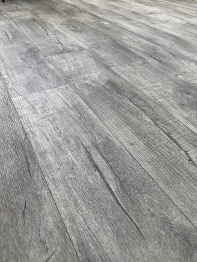 This is a close-up, angled shot of a gray wood-look floor. The planks are laid parallel to each other and run diagonally from the bottom left to the top right of the frame. The texture of the floor suggests it is laminate or vinyl flooring, mimicking the grain and imperfections of natural wood. The lighting creates subtle shadows along the seams between the planks, highlighting the horizontal grain pattern. There are no people, objects, or visible environmental features other than the flooring itself. The location within Tabuny, Russia, is not discernible from the image alone.