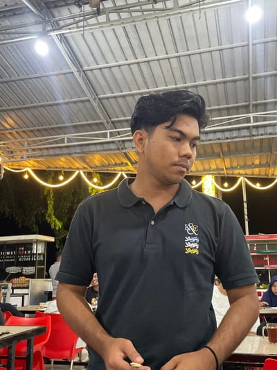 The image depicts a nighttime scene at a semi-outdoor dining establishment in Lhok Mereubo, Indonesia. The central subject is a young adult male, appearing to be of Southeast Asian ethnicity, positioned from the chest up and angled slightly to his right, with his gaze directed downwards and to the viewer's left. He has dark, neatly styled hair and wears a short-sleeved dark polo shirt featuring a multi-part crest with "K&C" text and four stylized lion figures (three white, one yellow) embroidered on the left chest. His right hand is visible, holding a small, light-colored, elongated object between his thumb and forefinger. A black string bracelet is present on his left wrist.

The environment features a light grey corrugated metal roof supported by white metal trusses overhead, illuminated by bright white circular LED light fixtures. Below the roof structure, against a dark sky, a string of warm yellow incandescent-style bulb lights is strung in draped festoons, with some green foliage dimly visible. In the background, multiple dark-topped tables and red plastic chairs are occupied by blurred patrons. To the far left, a white food stall or cart with indistinct lettering and hanging fabric is visible. To the far right, a female patron wearing a dark head covering sits at a table with a glass teacup in front of her. Another patron in the mid-left background appears to be smiling. The overall setting is a casual, artificially lit eatery.